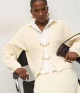 Cardigan de punto color crema con calce oversize, mangas amplias y cierre frontal mediante hebillas metálicas decorativas. Presenta una textura estructurada y puños ajustados.