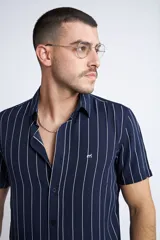 Camisa de manga corta con diseño a rayas verticales en color azul marino y blanco. Presenta cuello clásico, cartera abotonada frontal y logo bordado en el pecho. Posee un corte slim fit.