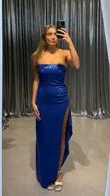 Vestido largo de fiesta color azul eléctrico, confeccionado en tela con acabado brillante. Presenta un diseño strapless con escote recto y una abertura lateral profunda en la falda.