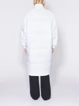 Plumífero blanco de corte recto y largo maxi, con cuello alto y cierre frontal con botones a presión. Incluye cinturón en la cintura.