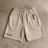 Bermuda de gabardina color beige con corte holgado. Presenta un bordado en tipografía gótica en la pierna derecha y un parche rectangular con el logo de la marca en la pierna izquierda. Cuenta con cintura elástica y bolsillos laterales.