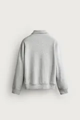 Sudadera gris de corte relaxed fit con cuello alto y cierre frontal de cremallera corta, confeccionada en algodón con interior de rizo afelpado. Presenta mangas largas, puños y bajo de rib, y un parche bordado con el logotipo de Fruit of the Loom en el pecho. Colección especial Fruit of the Loom x Zara.