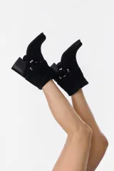 Bota de caña media confeccionada en cuero gamuzado color chocolate. Presenta un diseño de estilo western con taco cuadrado de 7 cm y una correa decorativa en el tobillo con herrajes metálicos plateados.