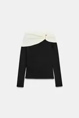 Top de manga larga con diseño bicolor, cuerpo negro y escote recto en color crema. Presenta un detalle de nudo con hebilla metálica dorada en el escote.