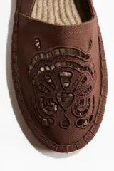 Alpargatas de lona de algodón en color marrón con detalles de broderie calados en la parte frontal y diseño bordado en el talón. Cuenta con suela de yute trenzado.