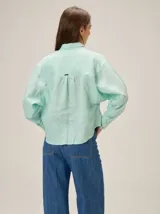 Camisa de lino color verde agua, con cuello clásico, cierre frontal de botones y mangas largas con puños. Presenta detalles de costuras en zigzag en la zona de los hombros y canesú.