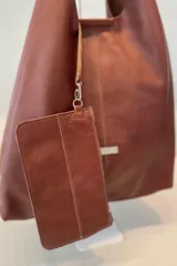 Bolso tipo tote de gran capacidad, confeccionado artesanalmente en cuero vacuno color marrón. Presenta un diseño minimalista con costura central vertical, fuelle lateral ajustable con cierre y placa metálica con logo en la parte inferior.