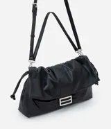 Cartera tipo shoulder bag de tamaño grande, confeccionada en poliuretano con acabado que imita el cuero. Presenta un diseño desestructurado con solapa superior, cierre mediante cordón de ajuste y detalles metálicos plateados en el frente. Incluye correa ajustable para llevar al hombro o cruzada.