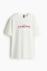 Remera de corte oversized en color blanco, con cuello redondo y mangas cortas de hombros caídos. Presenta un estampado gráfico central con la inscripción 'CORTINA' en color rojo y detalles en negro.