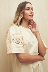 Vestido corto color crema, de corte recto y cuello redondo. Presenta mangas cortas amplias con calados geométricos en el ruedo.