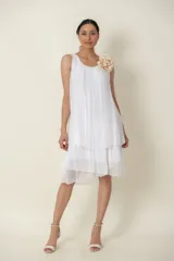 Vestido corto de seda blanca sin mangas, con diseño suelto, escote redondo y falda con volados escalonados. Incluye un detalle de flor de tela en el hombro.