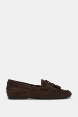 Mocasín de serraje color marrón, con detalle de fruncido en la puntera y lazo con borlas decorativas en el empeine. Presenta una suela plana de 1,5 cm.