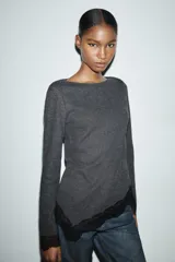 Top de manga larga en tejido suave color gris, con cuello redondo y bajo asimétrico decorado con encaje negro a tono. Presenta detalles de encaje negro en los puños.
