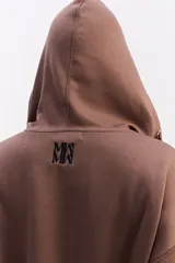 Hoodie de algodón color marrón con capucha, cierre frontal parcial y detalle de costuras horizontales en contraste negro. Presenta diseño holgado, mangas largas con puños elásticos y cordones ajustables en la capucha.