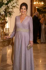 Vestido largo de lurex con acabado brillante en tono lila, con escote en V profundo, mangas largas y cintura marcada con un aplique bordado. La falda presenta un diseño drapeado que nace desde la cintura.