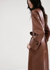 Chaqueta larga de efecto cuero color marrón, con cuello camisero, botonadura frontal oculta, bolsillos laterales y cinturón ajustable con hebilla en la cintura. Cuenta con trabillas en las muñecas y diseño amplio.