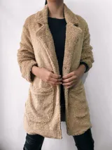Saco de abrigo color beige, de textura suave tipo peluche, con cuello solapa, cierre frontal con botones y bolsillos laterales.