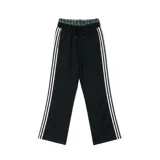 Pantalón deportivo negro de corte ancho, con tres franjas blancas verticales a lo largo de las piernas. La cintura es elástica y presenta un forro interior con estampado a cuadros en tonos verde y negro.