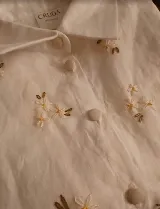 Camisa de lino 100% natural, color blanco, de silueta femenina y cómoda, con cuello clásico, cierre frontal con botones forrados y delicados bordados artesanales a mano con motivos florales.