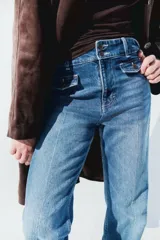 Jeans de denim de algodón con leve stretch, tiro alto, corte ajustado hasta la rodilla y acampanado desde la rodilla al bajo. Presenta cierre y botones frontales, y solapas de bolsillo decorativas con botón en la parte delantera.