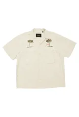 Camisa de manga corta color beige claro, estilo workwear/fatigue, con cuello camisero y dos bolsillos frontales con tapa y botón. Presenta bordados de palmeras en ambos lados del pecho y texto bordado en color naranja en el lado derecho.