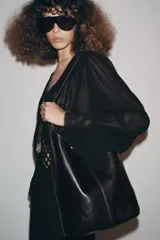 Bolso de hombro tipo saca asimétrica, color negro, con acabado de piel brillante. Posee un asa de hombro ajustable y cierre magnético. Incluye una cartera interior extraíble.