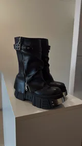 Botas de cuero negro con caña media, plataforma track y cierre de cremallera lateral. Presentan una correa con hebilla y ojales en la parte superior de la caña.