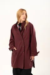 Trench de corte holgado color bordó, con cuello camisero, cierre frontal con botones y cinturón ajustable con hebilla. Presenta mangas largas con detalles de correas en los puños y bolsillos laterales.