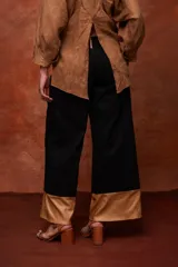 Pantalón ancho de tiro alto color beige, con pliegues frontales y botones visibles en la cintura. El ruedo del pantalón tiene un detalle de terminación en cuero color caramelo.