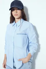 Camisa de silueta amplia y caída fluida, color celeste con rayas verticales blancas. Presenta cuello camisero clásico, cierre frontal con botones y un bolsillo aplicado en el pecho. Las costuras están resaltadas en azul oscuro.
