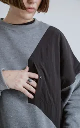 Buzo de cuello redondo con diseño oversize y corte crop. Confeccionado en frisa de algodón color gris melange, presenta un recorte asimétrico en color negro en microfibra con un bolsillo frontal funcional con cierre a presión.