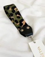 Llavero de tela con estampado animal print en tonos verde, negro y beige. Tiene un mosquetón metálico plateado y detalles en cuero sintético negro.