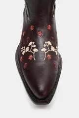 Bota estilo cowboy de cuero marrón oscuro con bordados florales en tonos claros y rojizos. Presenta tacón ancho en bloque, puntera afilada y tiradores laterales para facilitar el calce.