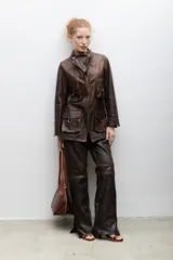 Blazer de cuero marrón con cierre frontal, cuello alto, múltiples bolsillos con hebillas y detalle de triángulo de cuero en el pecho.