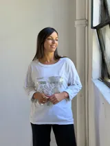 Remera blanca de manga larga con cuello redondo y diseño holgado. Presenta un aplique frontal rectangular con lentejuelas plateadas y detalles de encaje bordado.