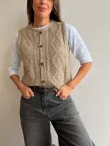 Chaleco tejido de punto color beige con diseño de rombos y trenzas en el frente. Presenta cuello redondo y cierre frontal con botones.