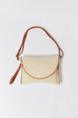 Bolso de mano tipo sobre confeccionado en rafia color crudo, con solapa de cierre y correa de cuero sintético marrón.