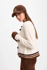 Cardigan de punto color crudo con detalles en marrón oscuro. Presenta cuello redondo, cierre frontal con botones tipo alamares y puños y ruedo acanalados con rayas horizontales. Incluye un pequeño aplique bordado en la manga.