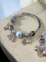 Pulsera rígida plateada con dijes colgantes y cuentas decorativas. Incluye cuentas de piedra veteada, cristales facetados en tono verde petróleo, una cuenta de cerámica blanca con diseño floral y múltiples dijes de corazón, uno de ellos con la frase 'you love you' grabada.