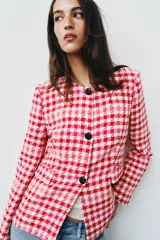 Blazer corto de tweed con estampado a cuadros en tonos rojo, rosado y blanco, cuello redondo, manga larga y cierre frontal con botones negros.