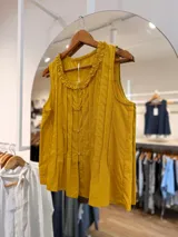 Blusa sin mangas de color mostaza, con tablitas verticales frontales, cierre de botones y detalle de volado fruncido en el cuello.