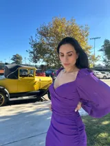 Vestido violeta con escote cuadrado, mangas largas abullonadas, corte ajustado y detalle drapeado con lazo en la cintura.
