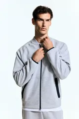 Campera bomber deportiva gris melange, de corte estándar, con cuello elevado corto, cierre bidireccional y mangas raglán. Posee bolsillos delanteros con cierre y costuras selladas, además de elástico fino en puños y bajo.