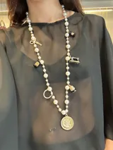 Collar largo de perlas blancas con dijes colgantes de metal dorado y esmalte, incluyendo motivos de costura como tijeras, carretes de hilo y una máquina de coser, finalizando en un dije circular central con diseño de flor.