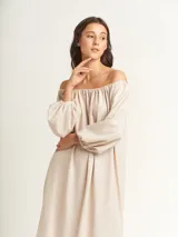 Vestido largo color beige, con escote elastizado que permite usarlo con los hombros descubiertos, mangas largas abullonadas, bolsillos laterales y tajos laterales en el ruedo.