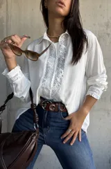 Camisa blanca de corte clásico con detalles de encaje floral vertical en la parte frontal y en los puños. Presenta cuello camisero, cierre frontal con botones y mangas largas con terminación elástica.