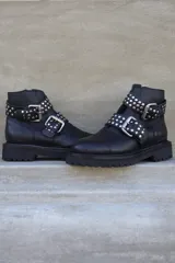 Borcego negro de cuero con plataforma y suela track. Presenta dos correas con hebillas plateadas y apliques de tachas redondas.