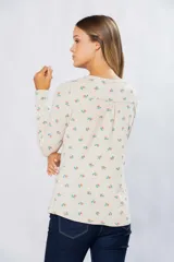 Remera de manga larga color crudo con estampado de flores pequeñas en tonos naranja y verde. Tiene cuello redondo y pin metálico con logo personalizado en el ruedo.