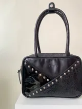 Cartera rígida negra con detalles en rojo y tachas plateadas. Tiene herrajes y cierre metálico plateado marca YKK, asa fina para llevar al hombro o en la mano y correa larga extra regulable. El interior es forrado, tiene un bolsillo con cierre enmarcado en cuero, y un segundo bolsillo plaqué.