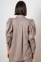 Camisa de algodón color marrón con mangas largas abullonadas, cuello clásico y cierre frontal oculto. Presenta botones decorativos con strass en los puños y en el cuello.